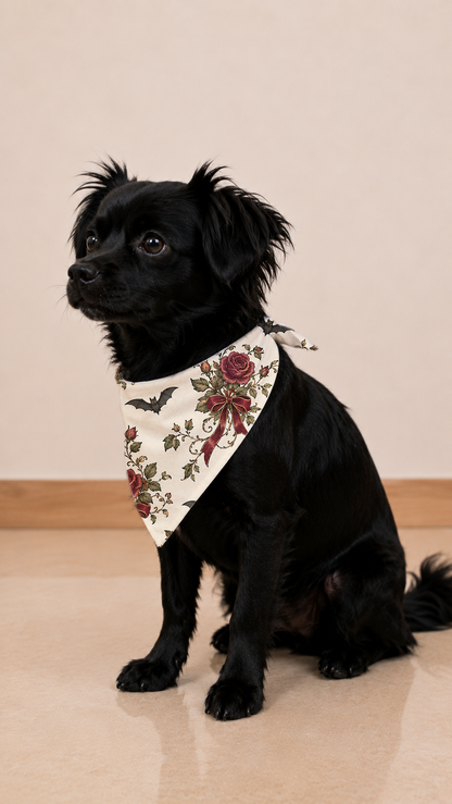A small black dog wearing a cream bandana with a red rose and bat pattern, sitting on a light beige tiled floor against a beige wall.