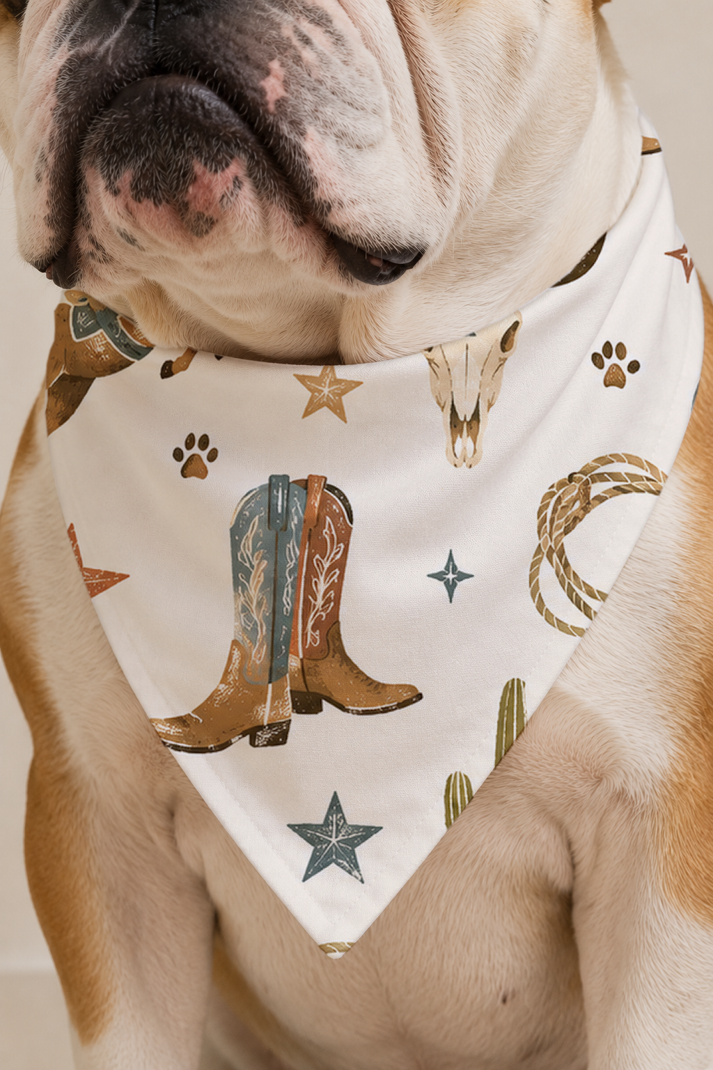 A close-up of a light-colored dog wearing a white bandana with a western print featuring cowboy boots, stars, paw prints, and lasso details.