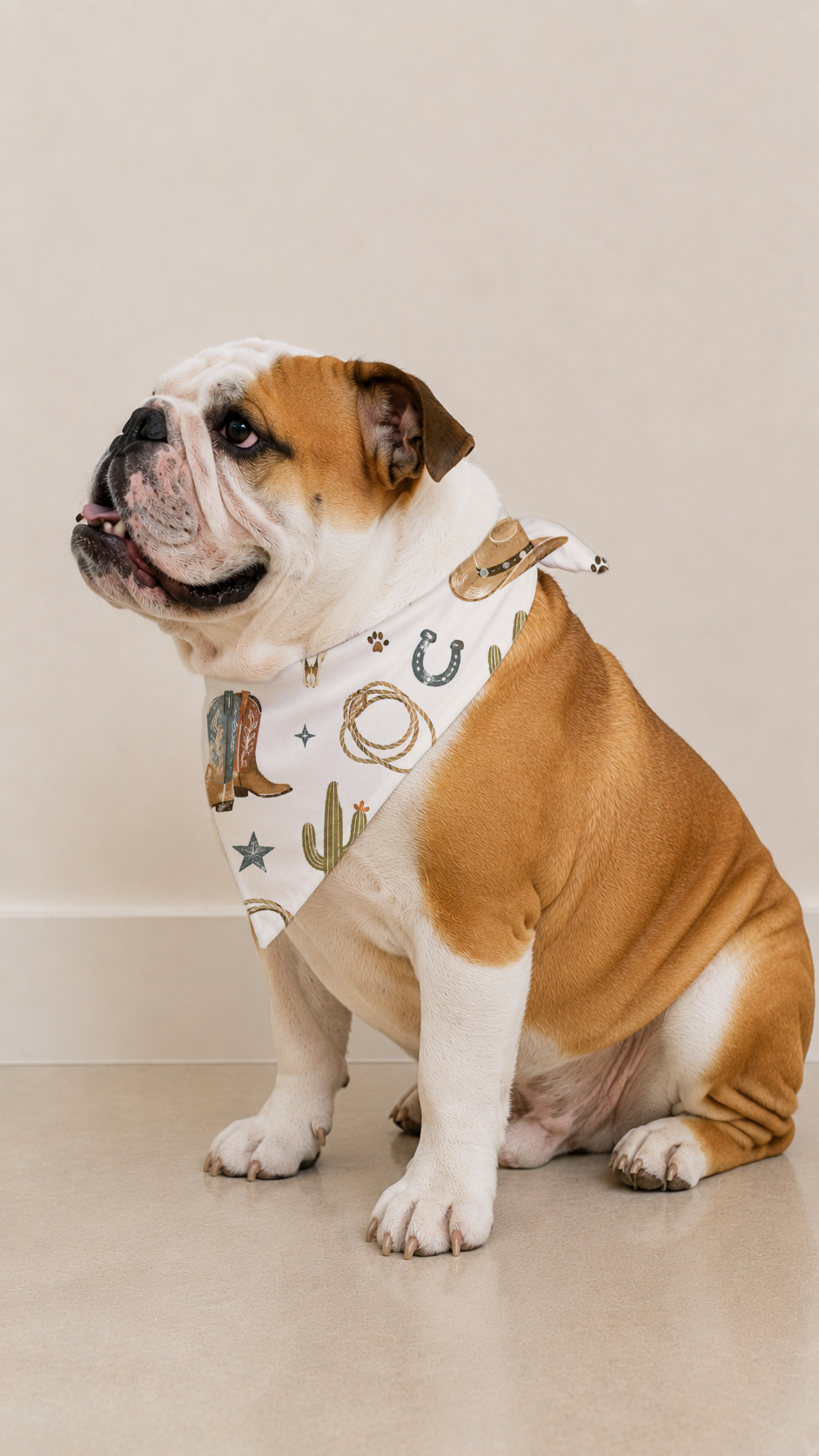 An English Bulldog wearing a white bandana with a western-themed print of boots, horseshoes, and cacti, sitting against a plain beige wall.